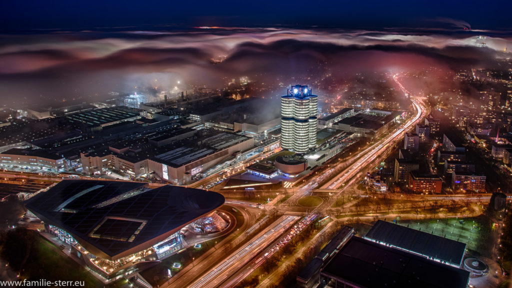 Eine Nebelwolke umhüllt das nächtlich beleuchtete BMW Hochhaus und die BMW - Welt