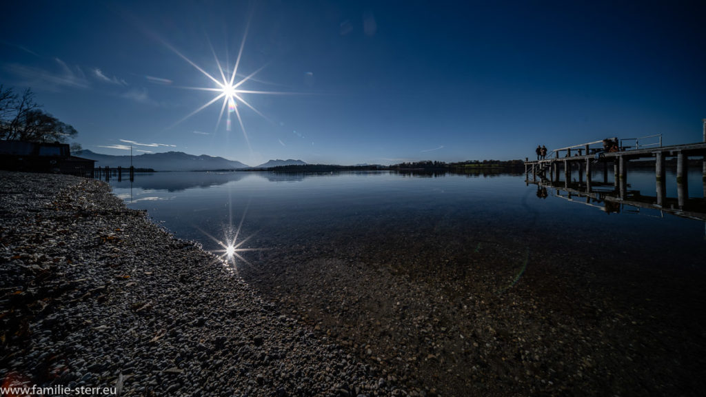 strahlende Sonne und blauer Himmel über dem spiegelglatten Chiemsee am Strandbad Breitbrunn auf der Halbinsel Urfahrn