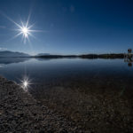 strahlende Sonne und blauer Himmel über dem spiegelglatten Chiemsee am Strandbad Breitbrunn auf der Halbinsel Urfahrn