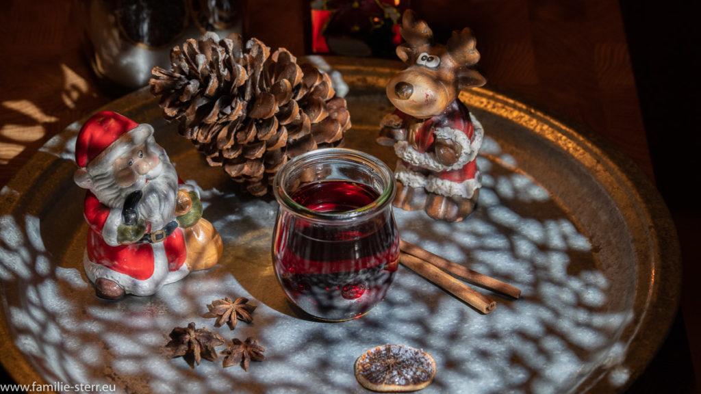 ein Glas Glühwein auf einem weihnachtlich dekorierten Teller mit Weihnachtsmann, Rentier, Tannenzapfen, Sternanis und Zimtstangen