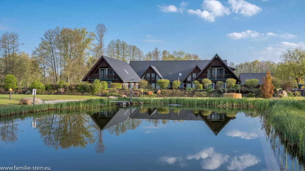 drei Holzhäuser in Form eines Spreewälder Dreiseitenhofs spiegeln sich im Naturbadeteich des Seehotels Burg Spreewald