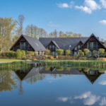 drei Holzhäuser in Form eines Spreewälder Dreiseitenhofs spiegeln sich im Naturbadeteich des Seehotels Burg Spreewald