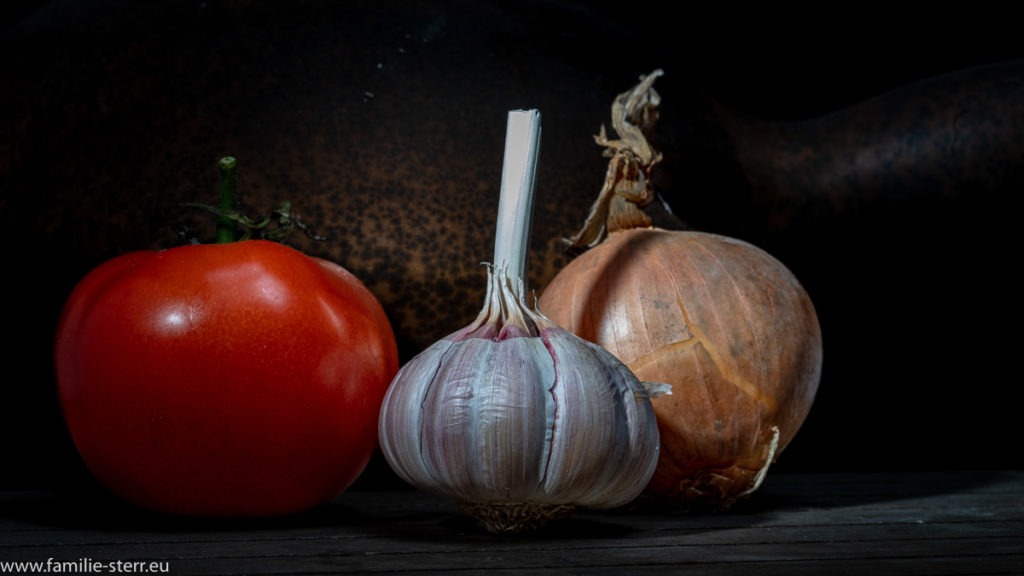 eine rote Rispentomate, eine Zwiebel und eine Knolle Knoblauch - die Zutaten für ein Bruschetta