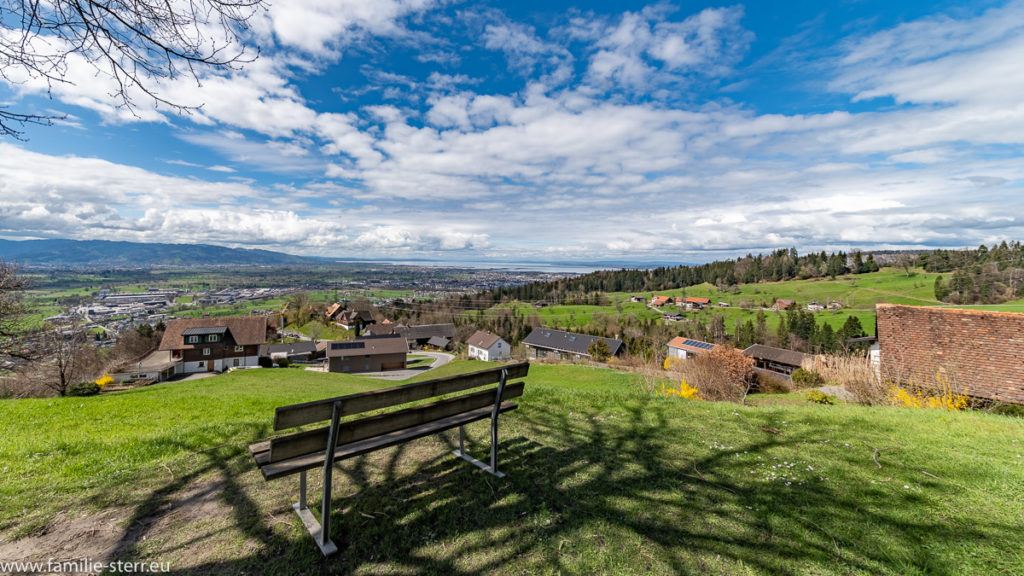 Bänkchen im Park vor der Basilika Maria Bildstein mit Blick über Bregenz und den Bodensee unter weiß-blauem Himmel