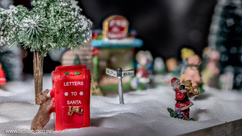 ein Hund sitzt vor einem Briefkasten für Weihnachtspost in einer schneebedeckten Landschaft / unser Modell - Weihnachtsdorf 2025, das Weihnachtsdorf ist aufgebaut auf weißem Kerzensand