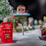 ein Hund sitzt vor einem Briefkasten für Weihnachtspost in einer schneebedeckten Landschaft / unser Modell - Weihnachtsdorf 2025, das Weihnachtsdorf ist aufgebaut auf weißem Kerzensand